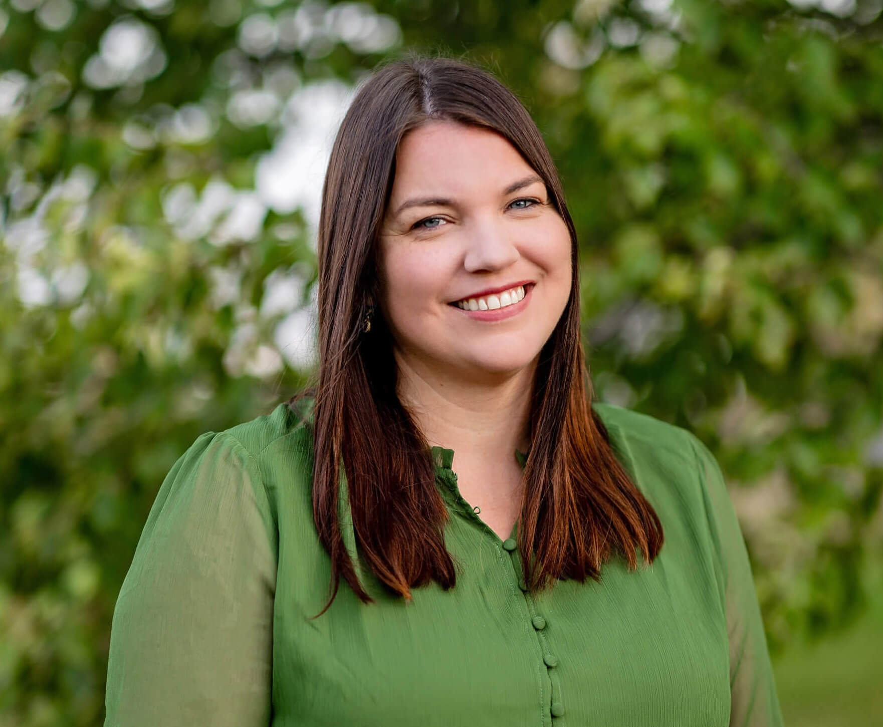 Sarah Casey is wearing a green shirt and is standing in front of a green tree and smiling.