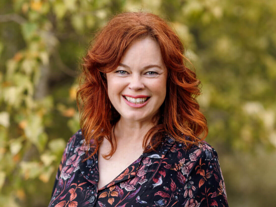 Lisa Turner is looking at the camera and smiling. She has bright red hair and is wearing a mulitcolored dress that has a black background. Lisa is standing in front of trees with green leaves.