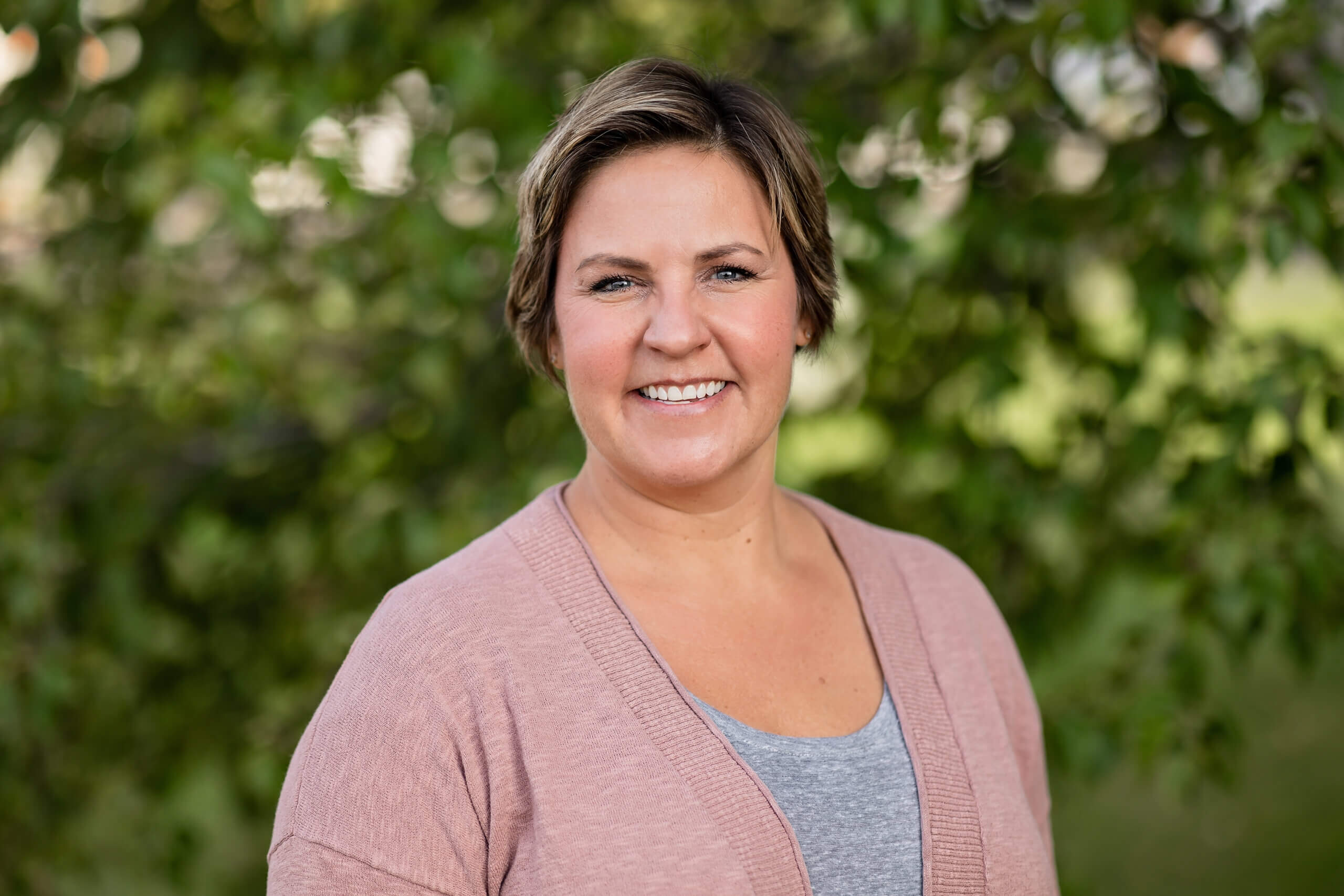 Laura has short brown hair and is wearing a pink cardigan and gray shirt. She is standing in front a tree with green leaves and is smiling.