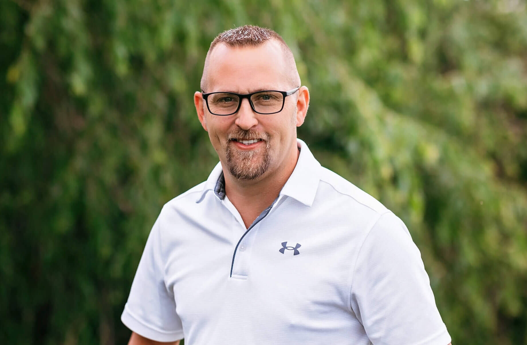Chad is standing outside in front of a tree. It is a sunny day. He is wearing glasses and a white shirt. He is smiling.