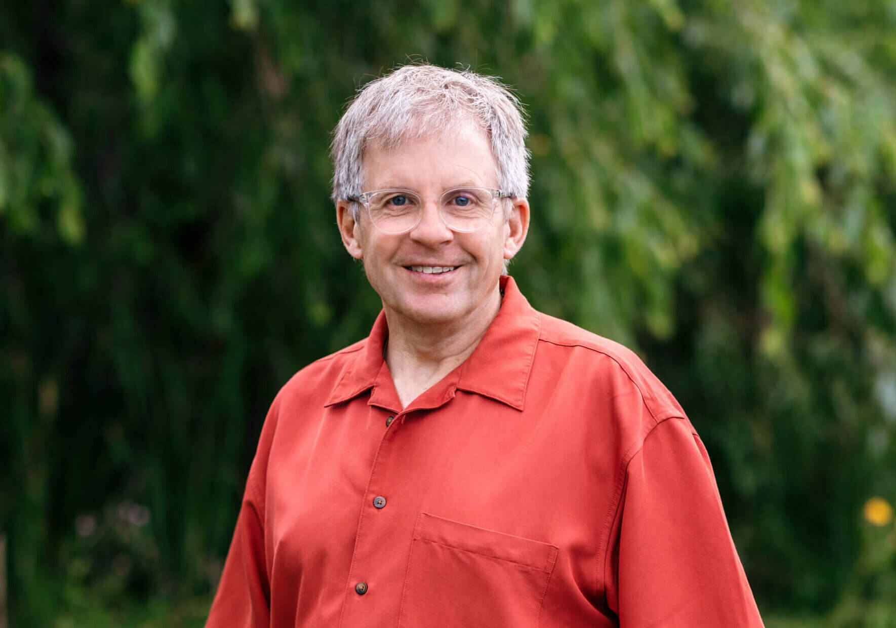 Kevin is wearing a bright orange-ish red shirt and glasses. He has short gray hair and is smiling. Kevin is standing in front of a green tree on a sunny day.