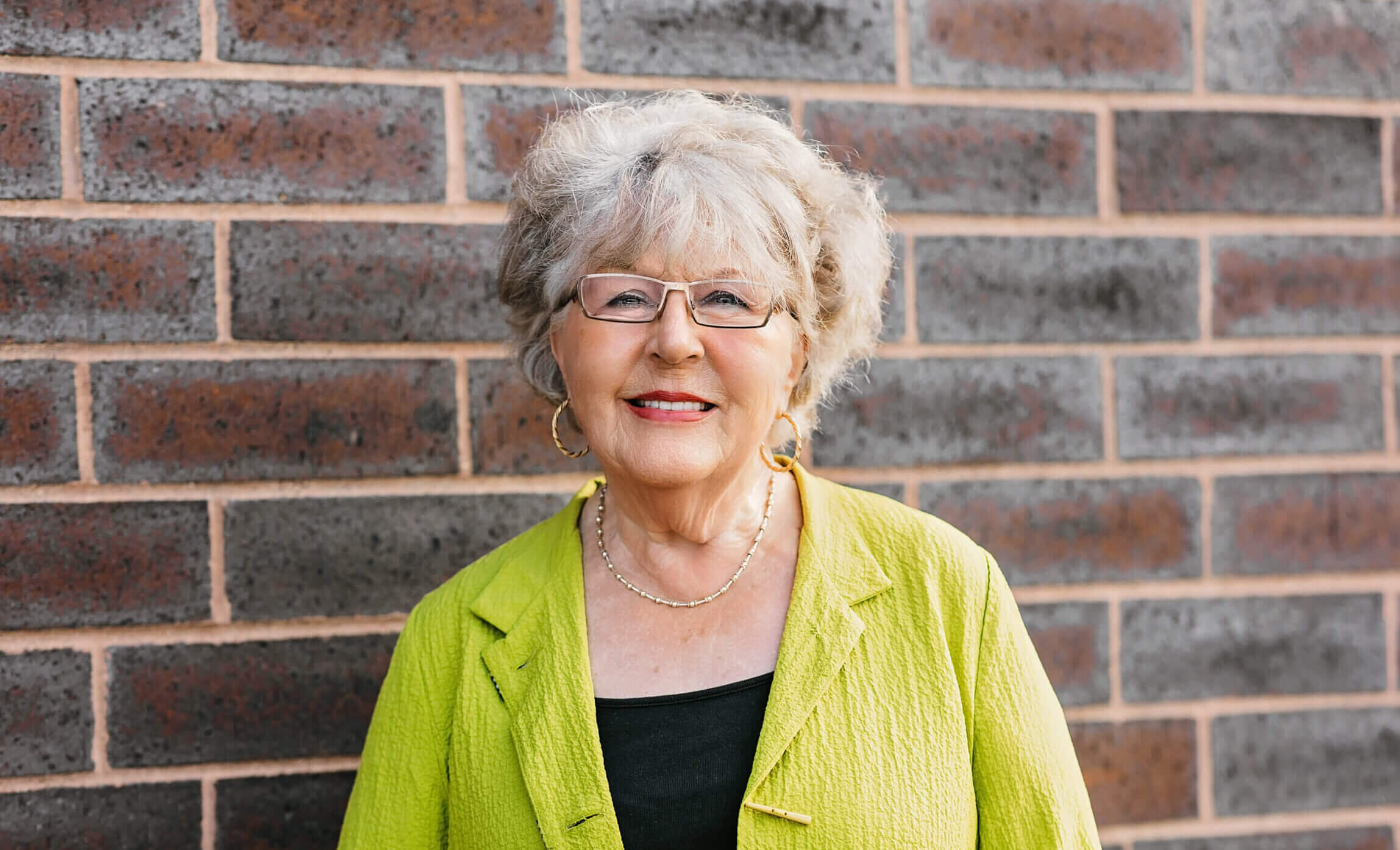 Patricia is wearing a bright green shirt. She is standing in front of a brick wall and smiling.