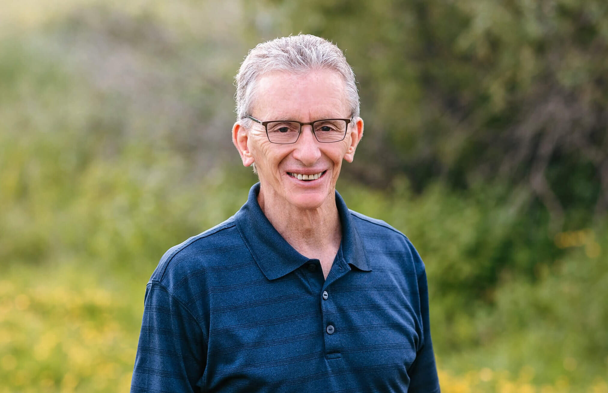 Paul is standing outside on a sunny day. He is wearing a blue shirt and glasses.