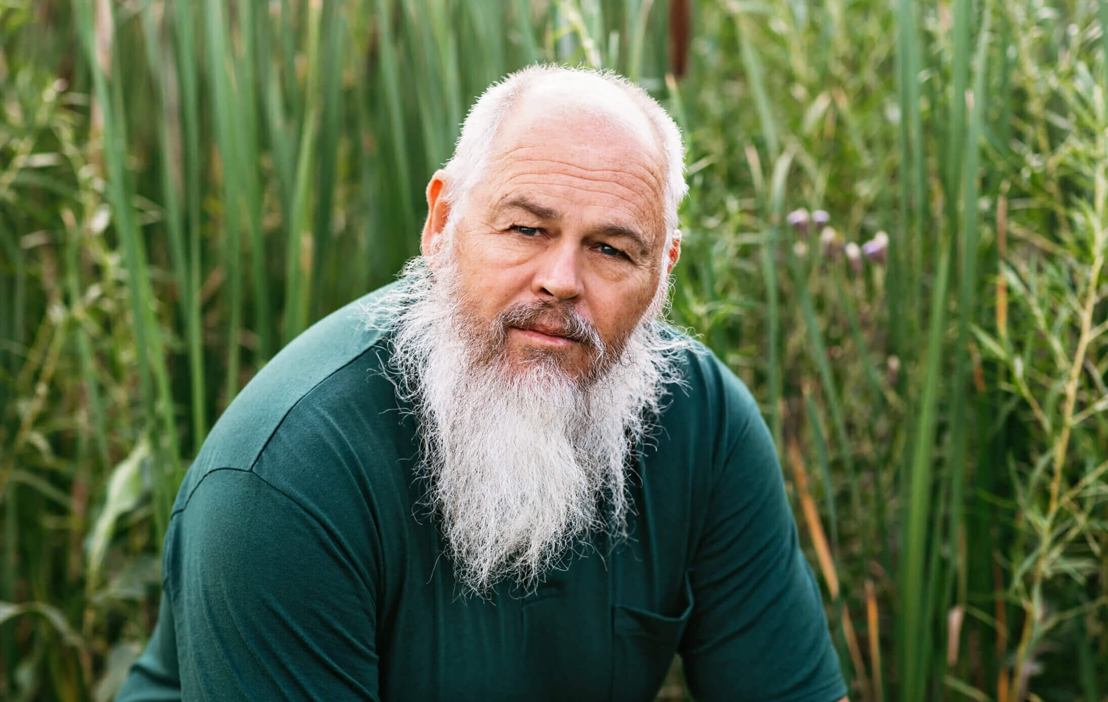 Paul is kneeling in front of green cattails. Paul is wearing a dark green shirt and has a long white beard.