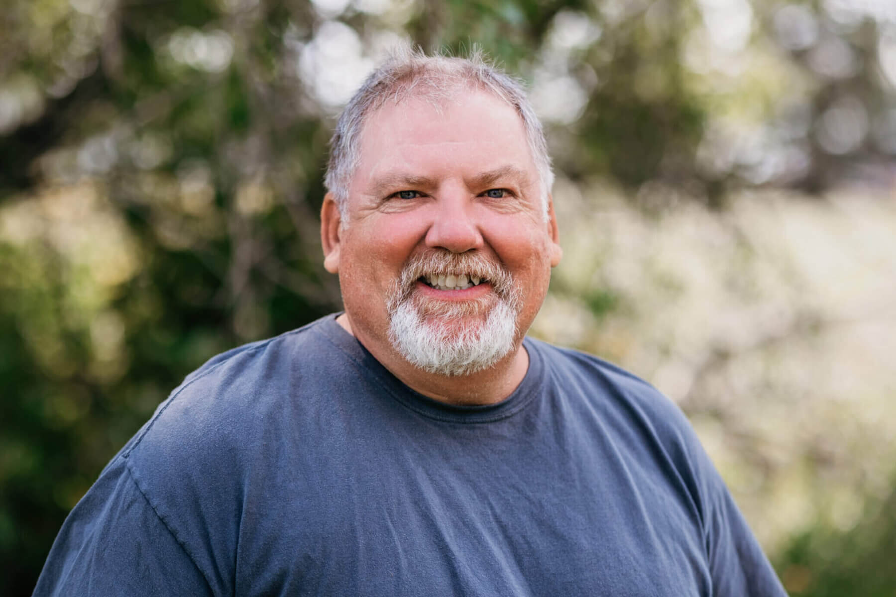 Rick is smiling and standing in front of a tree on a sunny day.