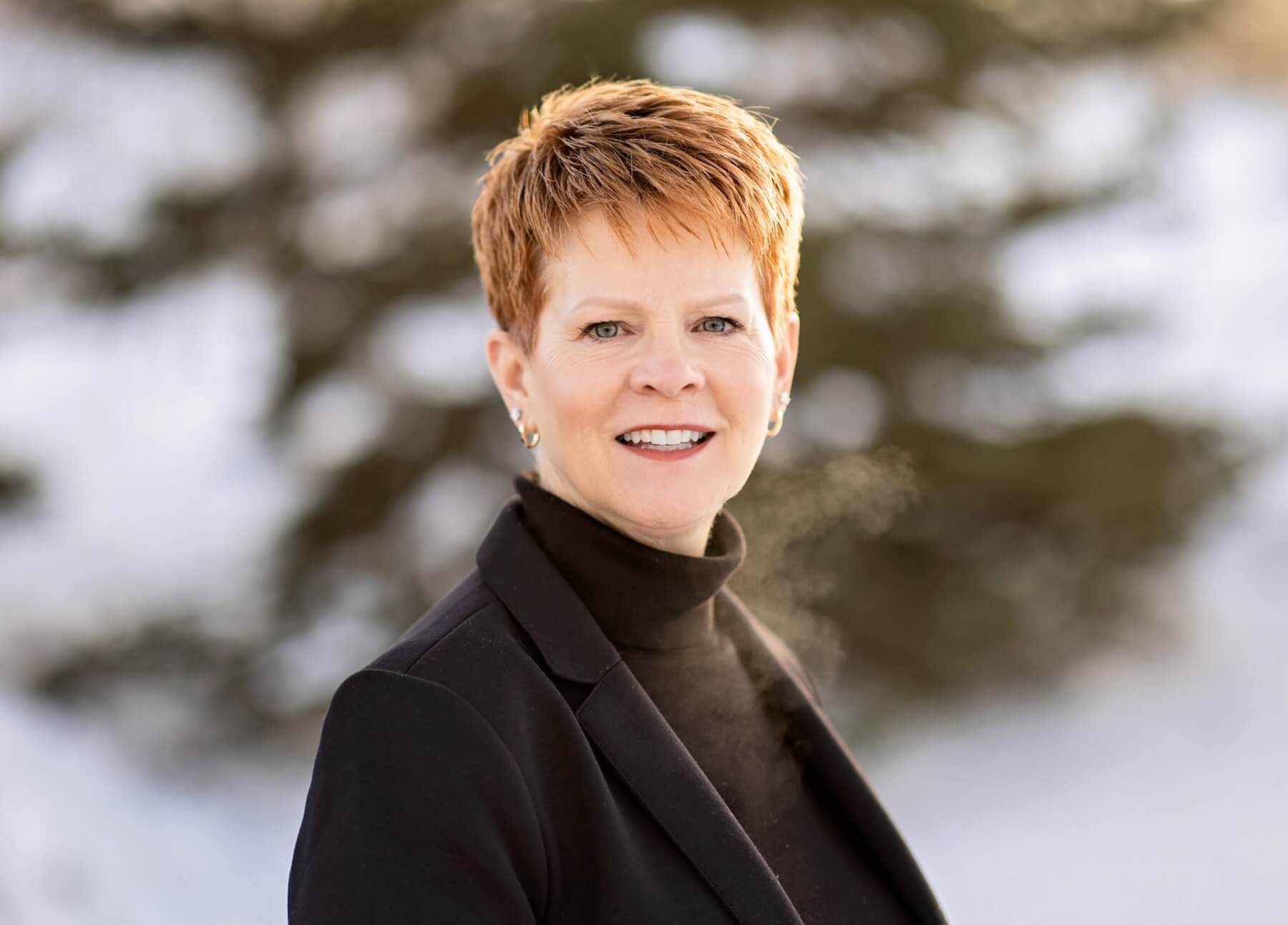 Jackie is standing outside on a winter day. She is wearing a back blazer and black turtle neck. She has short red hair. Jackie is facing the camera and smiling.