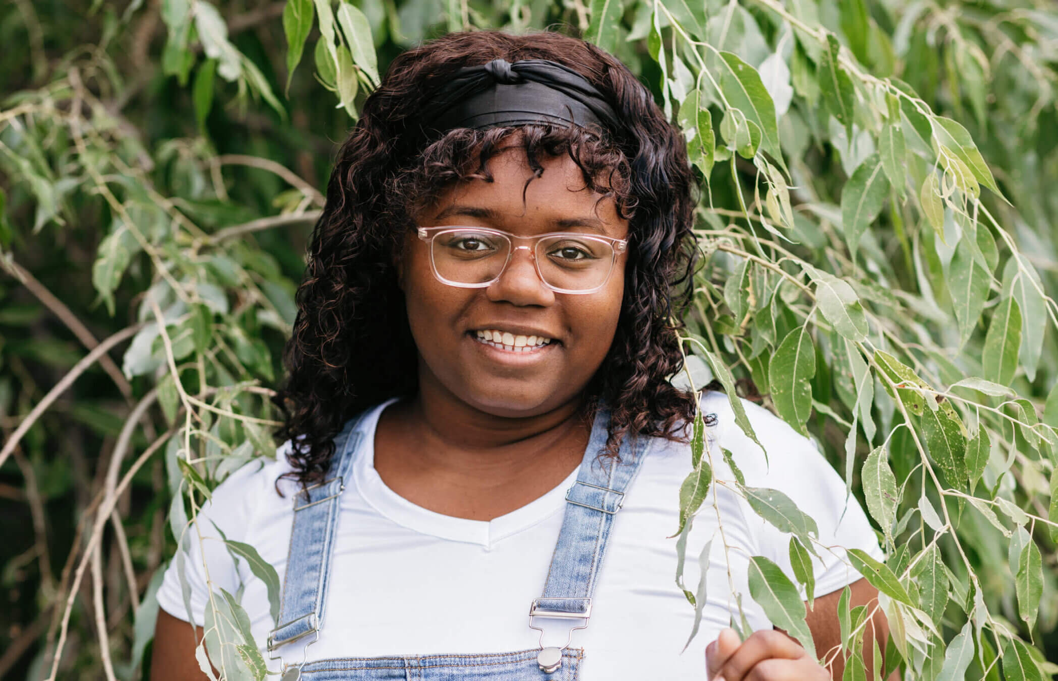 Aaliyah is standing amongst the branches of a tree, smiling. She is wearing overalls, a white shirt, glasses, and a black headband.