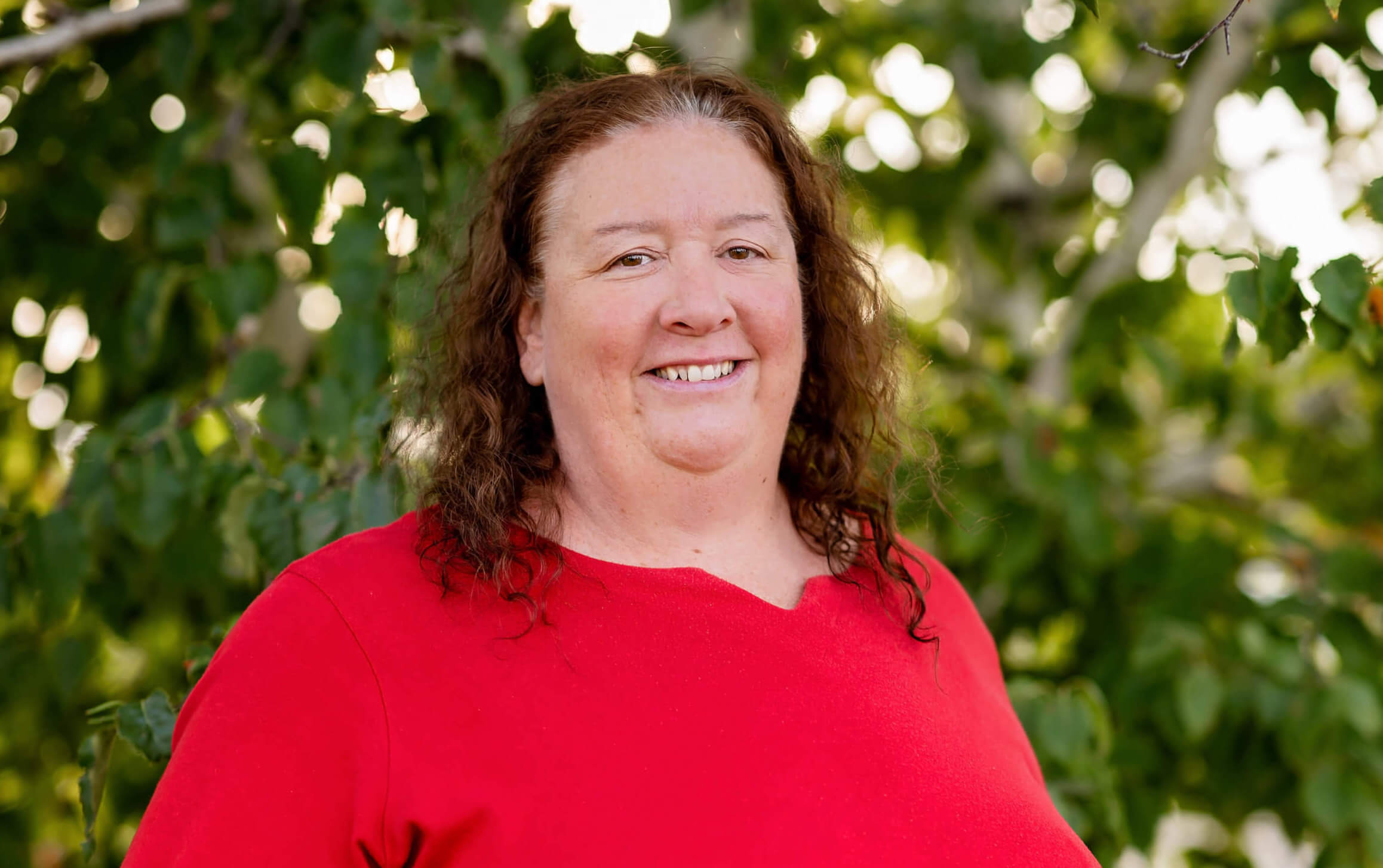 Michele is wearing a bright red shirt. She has brown hair and is smiling. She is standing in front of a tree with green leaves.