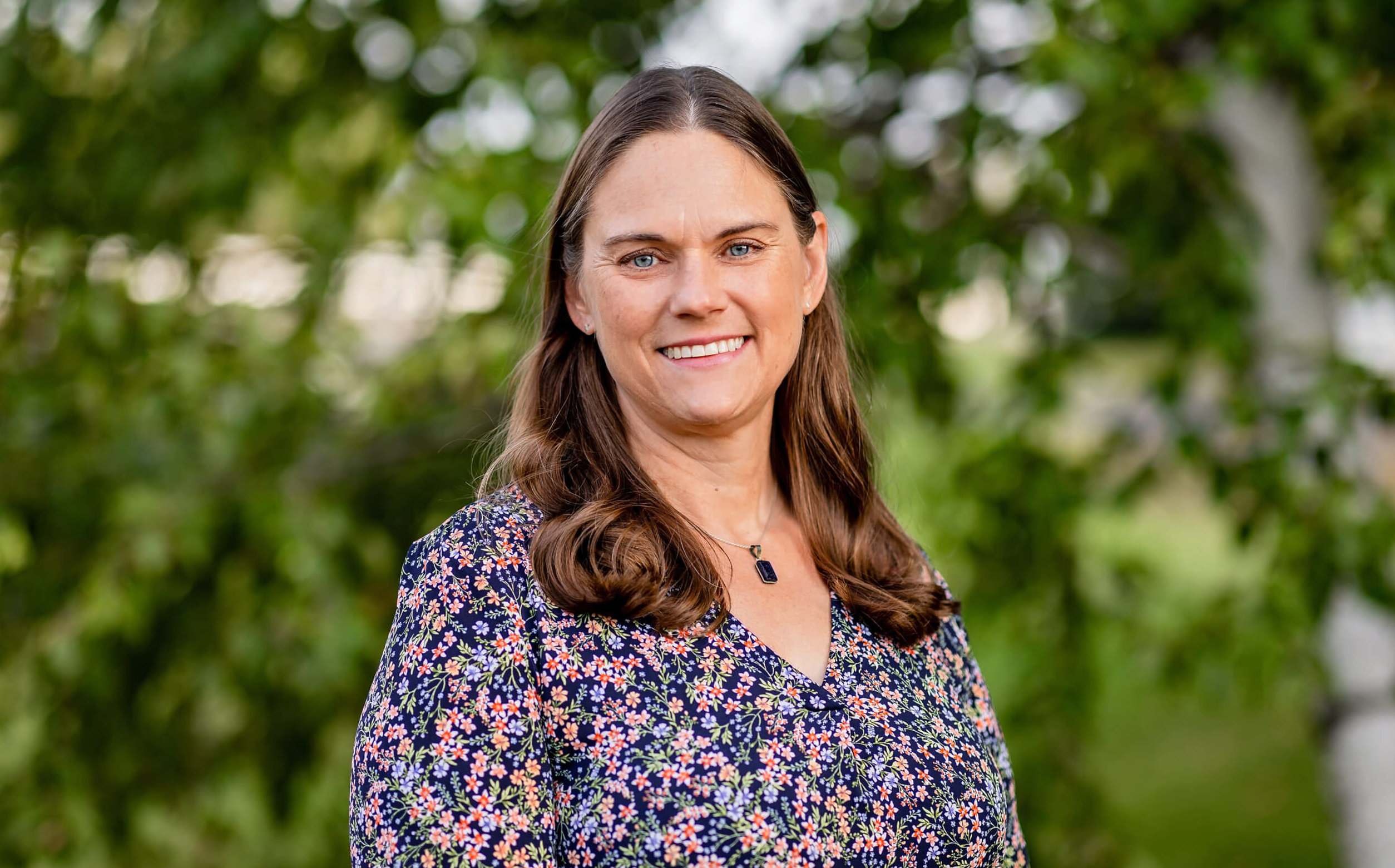 Trina Starker has long brown hair and is wearing a floral shirt. She is smiling and standing in front of a green tree.
