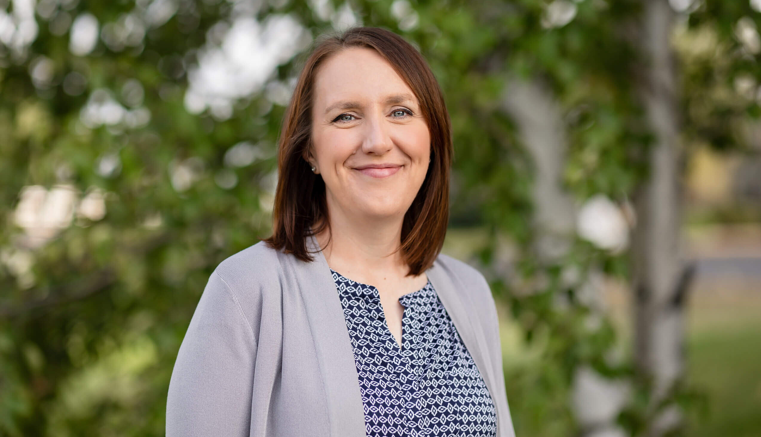 Janice is wearing a blue shirt and gray cardigan. She has shoulder length brown hair. She is standing in front of a birch tree with green leaves. Janice is smiling.
