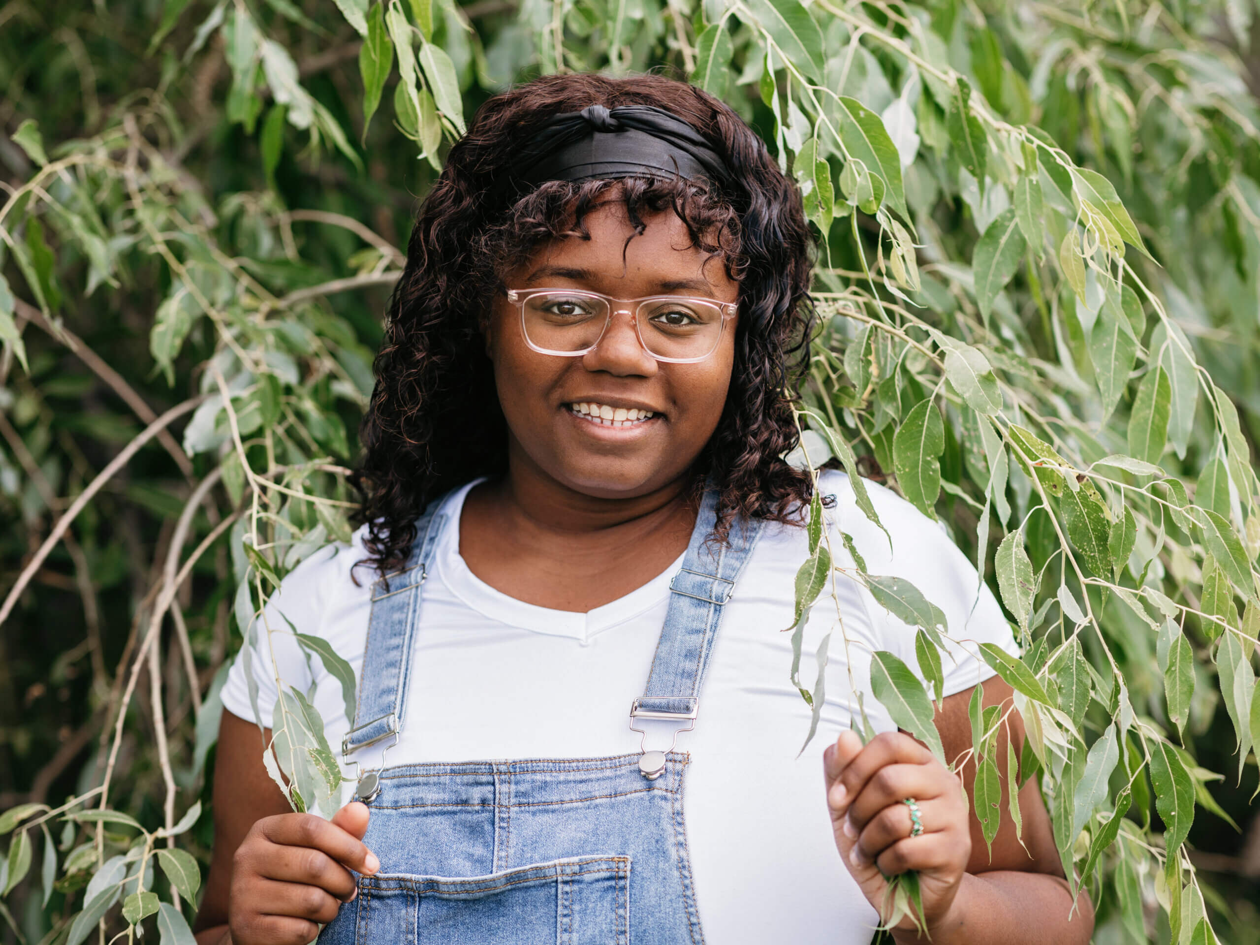 Aaliyah is standing amongst the branches of a tree, smiling. She is wearing overalls, a white shirt, glasses, and a black headband.