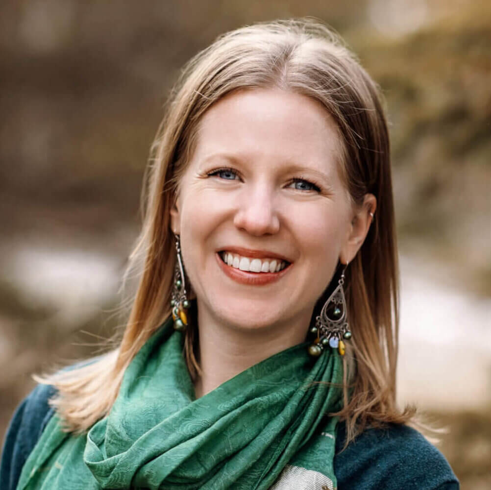 Amanda is standing in front of evergreen trees. She is smiling and wearing a green shirt and green scarf.