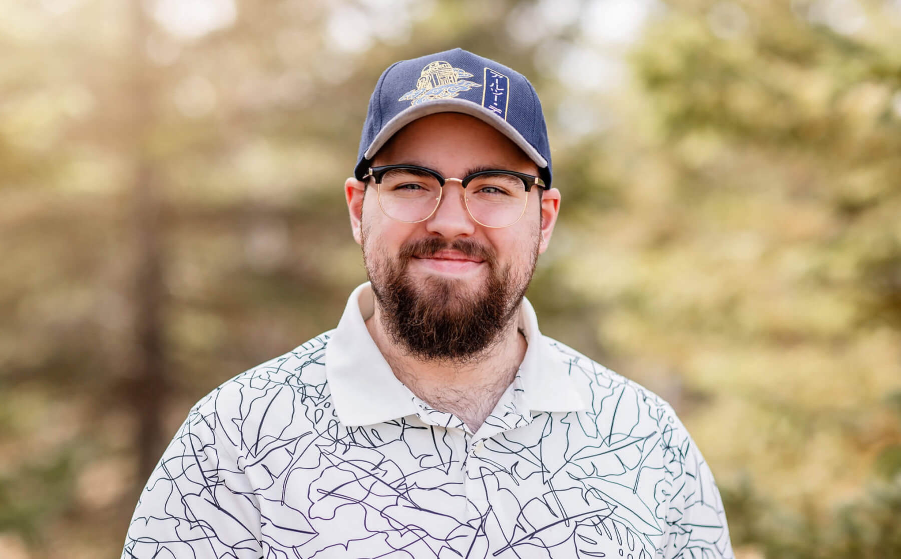John is standing in a wooded area on a spring day. He is wearing a white polo shirt, baseball hat, and glasses. He has a brown beard and is smiling.