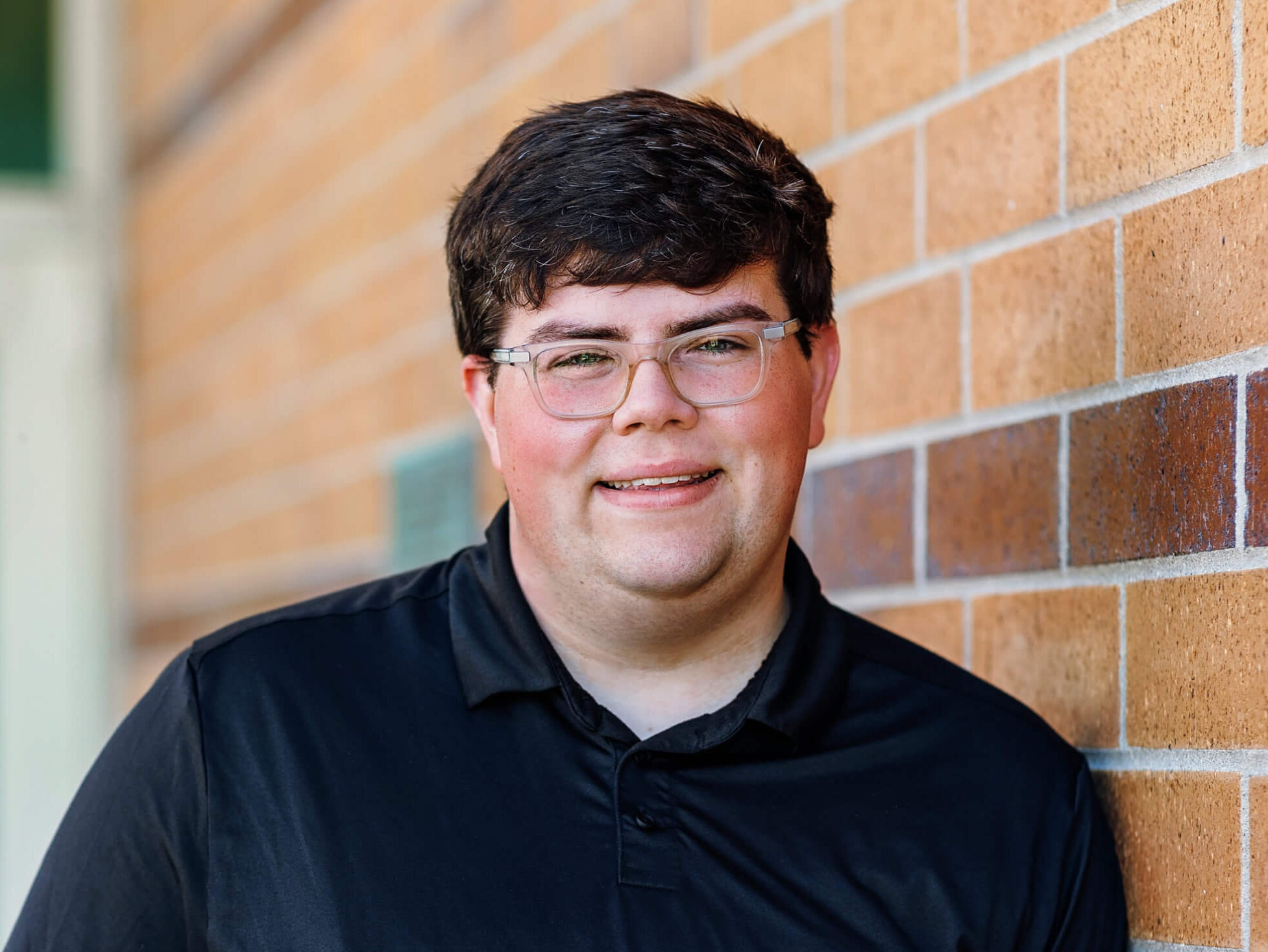 Griffin is wearing a black polo shirt and clear glasses. They have short brown hair. Griffin is smiling and leaning against a red brick wall.