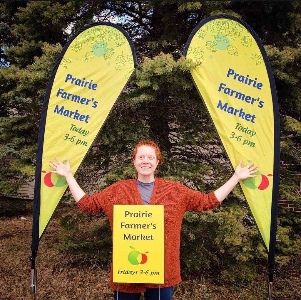 Picture of Meghan Janke smiling in-between to signs for the Prairie Farmer's Market