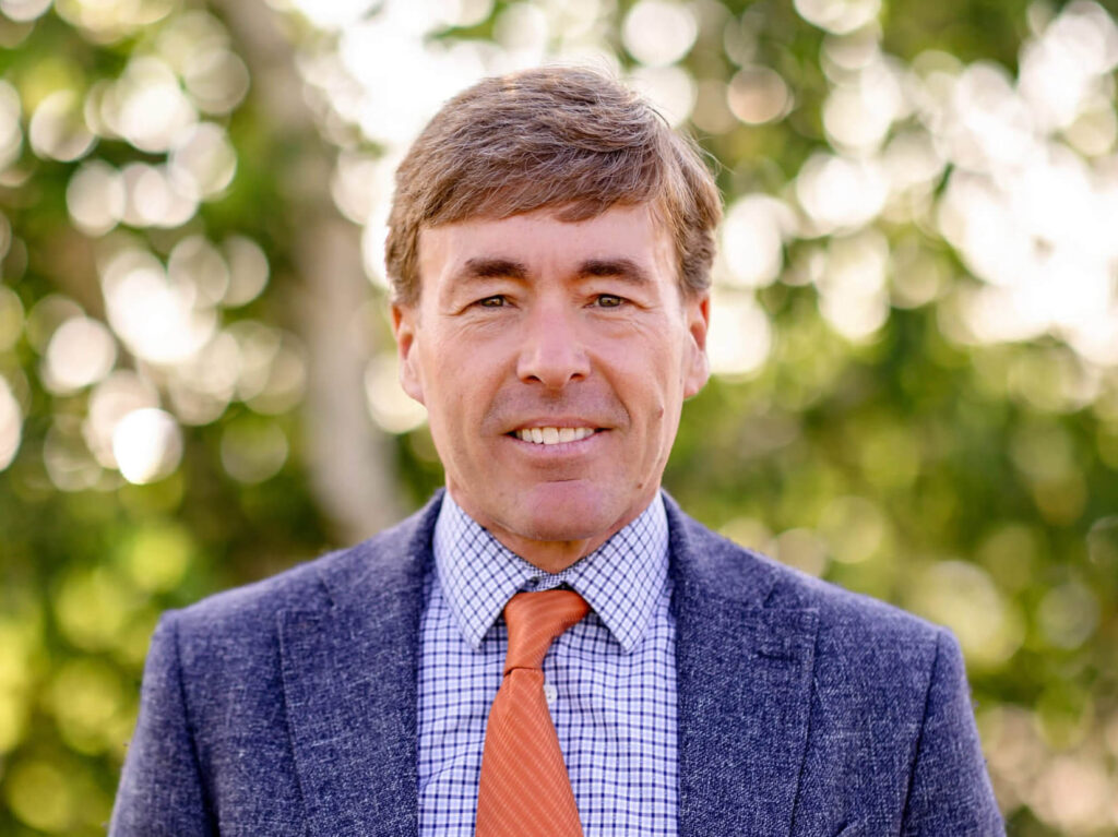 Ben Schierer is wearing a navy blue suit, light blue shirt, and orange tie. He is standing in front of green trees and smiling.