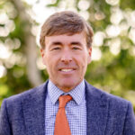 Ben Schierer is wearing a navy blue suit, light blue shirt, and orange tie. He is standing in front of green trees and smiling.