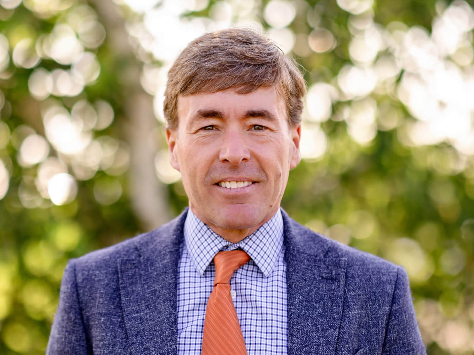 Ben Schierer is wearing a navy blue suit, light blue shirt, and orange tie. He is standing in front of green trees and smiling.
