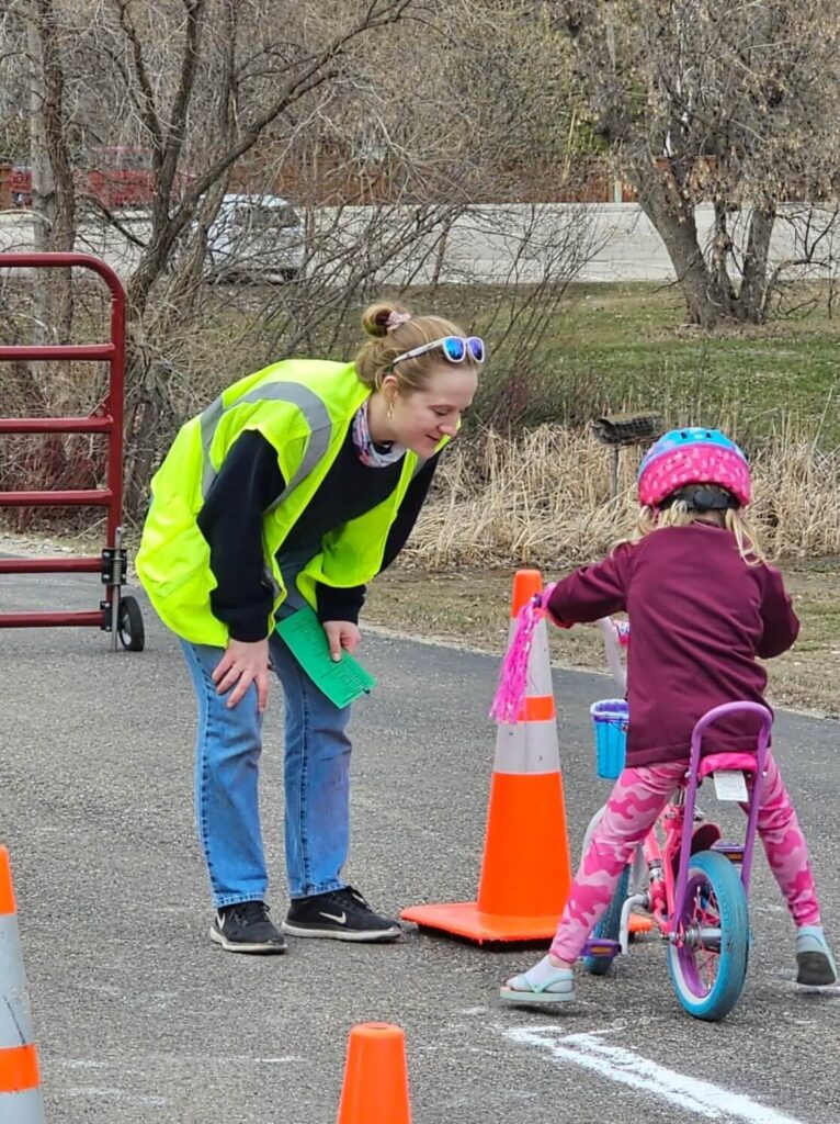 Mallory Jarvi is wearing a bright green traffic vest and helping a child wearing pink pants, shirt, and helmet on a pink bike.