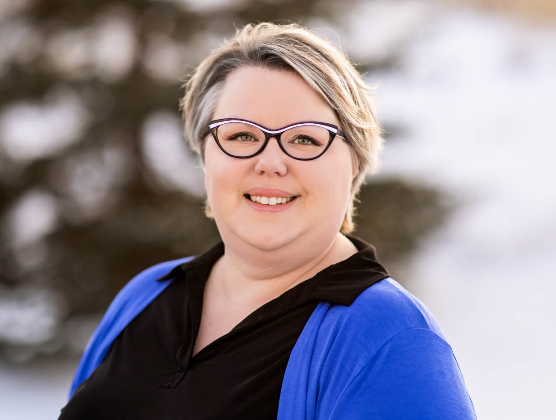 Lisa is standing outside on a winter day. She is wearing a royal blue cardigan and a black button up shirt. She has short light hair and is wearing glasses. She is facing the camera and smiling.