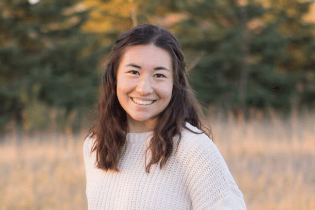 Isabel is smiling at the camera. She has brown hair and is wearing a white sweater.