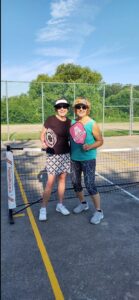 Two women in athletic outfits are smiling at the camera on a gray pickleball court. They are holding pickleball paddles and wearing visors and sunglasses. There is a net and sunlight in the background.