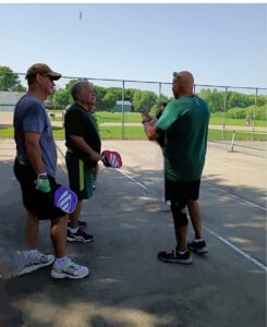 Three men are standing on a gray pickleball court. They are wearing athletic shorts, t-shirts, and tennis shoes. They are all holding pickleall paddles and talking.