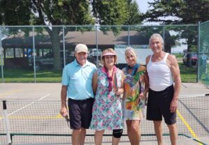 Two men in shorts and a T-shirt and two women in tennis dresses are smiling at the camera. They are standing on a gray pickleball court.