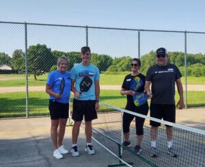Two women and two men are standing smiling on a gray pickleball court. Every person is holding a pickleball paddle. They are all wearing shades of blue and gray. They are in tennis shoes.