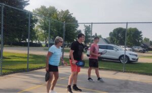 Three people are playing pickleball on a gray pickleball court. Everyone is holding a pickleball paddle. Ther is a grandma and two grandsons.