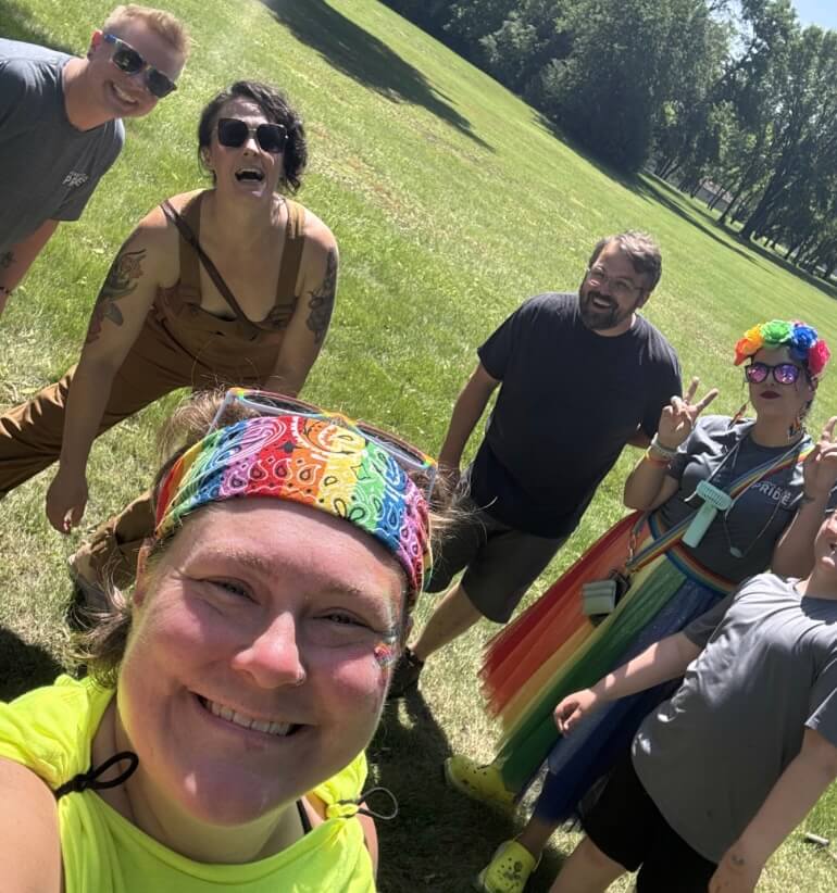 Six people are standing on the grass, smiling at the camera. Celebrating Pride.