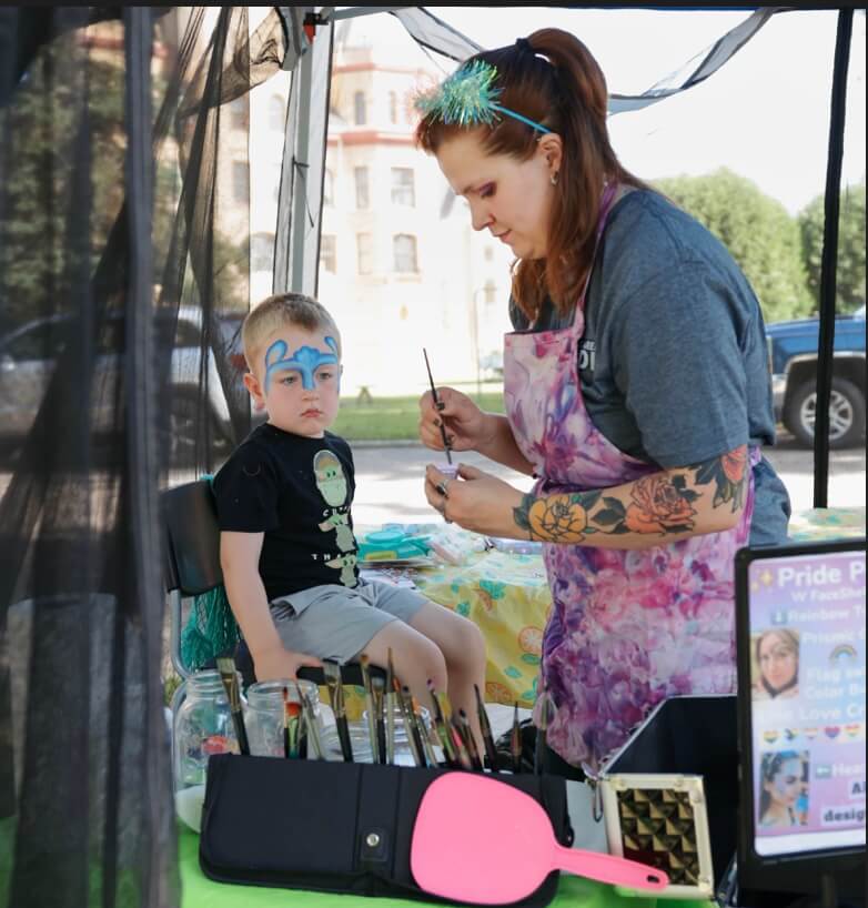 A woman in a pink and purple apron, gray t-shirt, and a blue headband is painting a little girl's face. The little girl is sitting in a colorful tutu and a black t-shirt.t