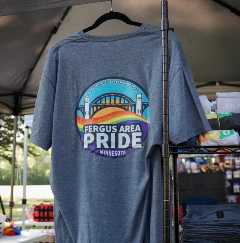A gray t-shirt is hanging on a black hanger in a tent. On the back is a colorful circle with a bridge and Fergus Area Pride written on it.