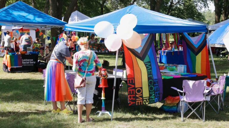 There are tent booths at Fergus Pride. The front of one tent has a Pride flag and white balloons. Two women, one in a rainbow skirt and one in a colorful shirt and white pants, stand under the tent celebrating.