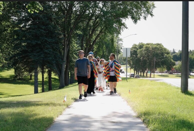 People in Pride colors march down a gray sidewalk to celebrate. Green trees and grass surround the sidewalk.