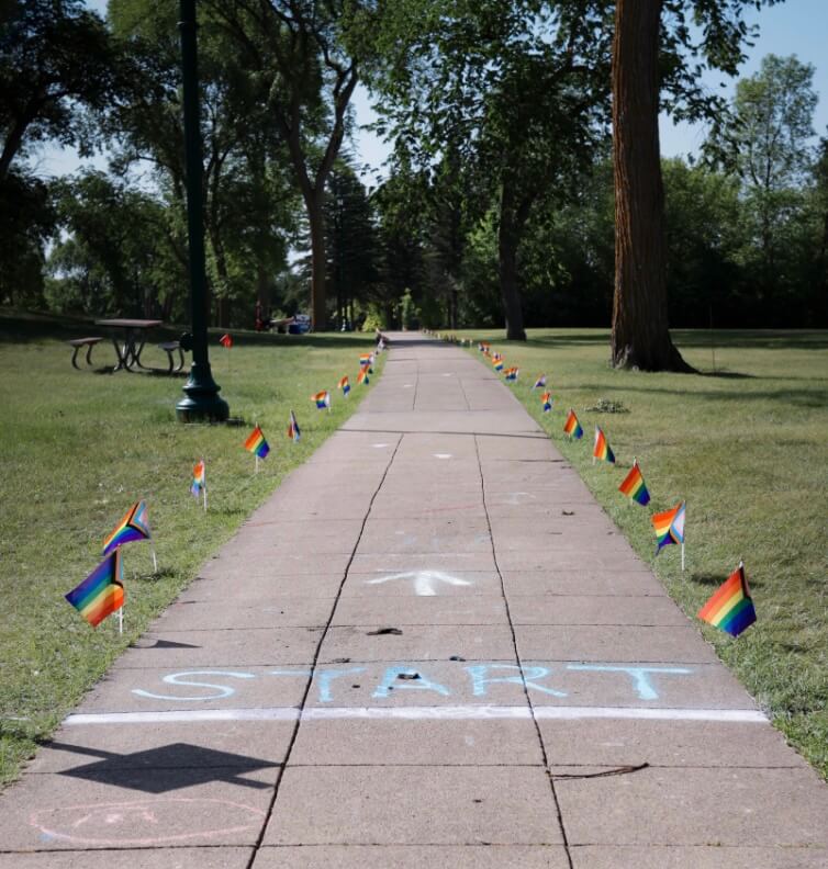 There is a start line and arrow on the gray sidewalk in blue and white chalk. The sidewalk is lined with little Pride flags. Trees surround the sidewalk.
