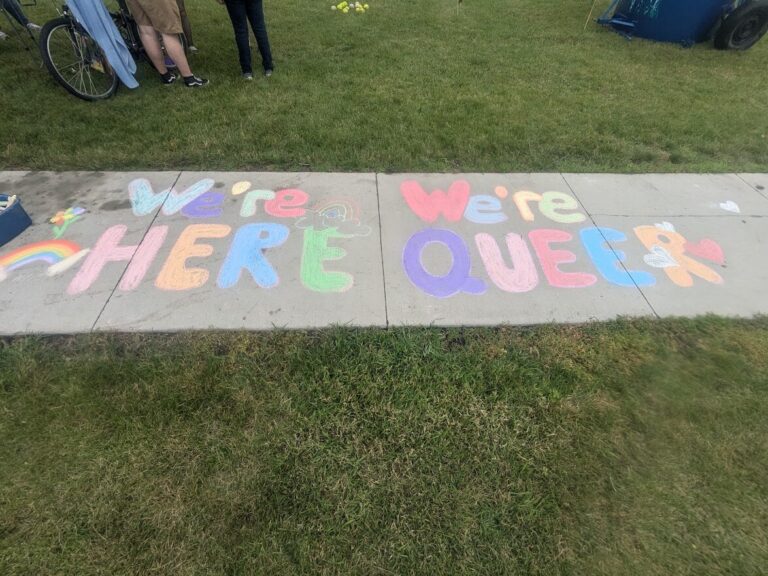On the gray sidewalk in large, colorful chalk letters spells We're here, We're queer.