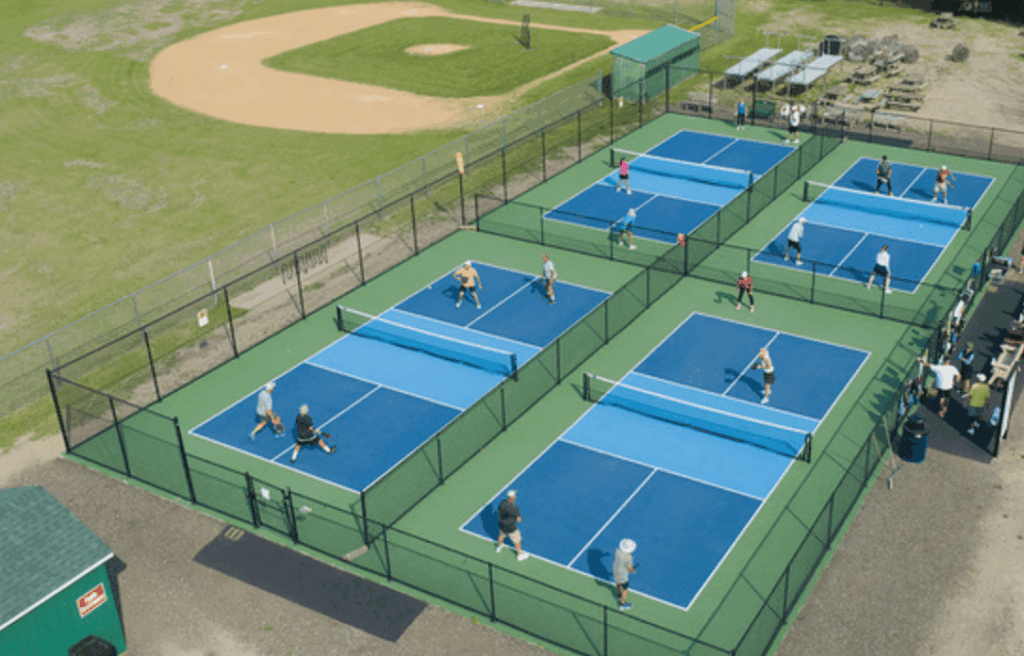 This image is taken from above. It shows four blue and green pickleball courts surrounded by a black chain link fence. There are people playing pickleball on the courts.