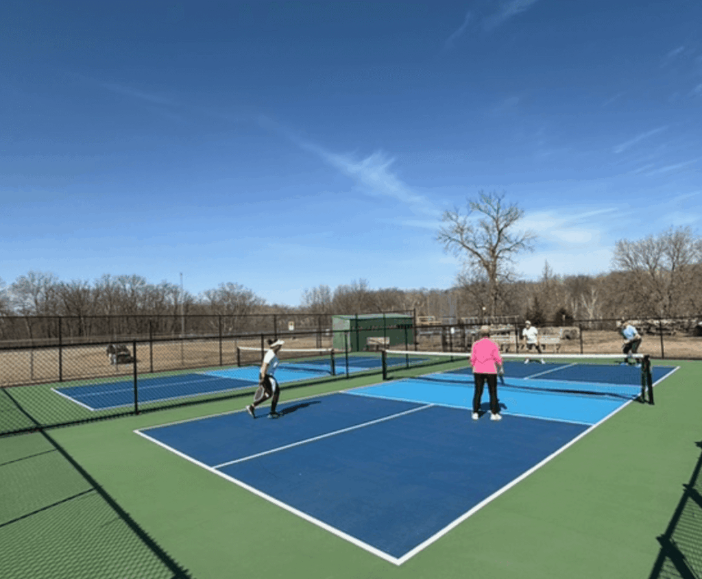 People are playing pickleball on dark blue courts. Each is holding a pickleball paddle. it is a sunny day.