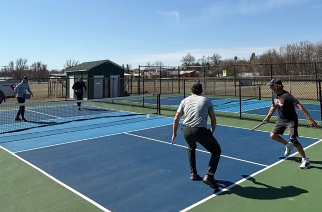 There are four men playing pickleball on a dark blue court. There is a net and chain link fence surrounding them.