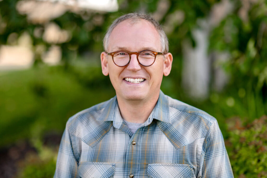 Justin is smiling. He's wearing brown round glasses and a blue and white plaid shirt. He stands in front of a green tree on a sunny day.