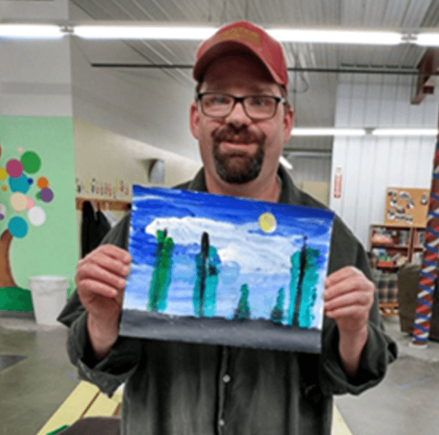 A man in a gray/green shirt and a red baseball hat is smiling at the camera and holding up a painting of trees and the night sky.
