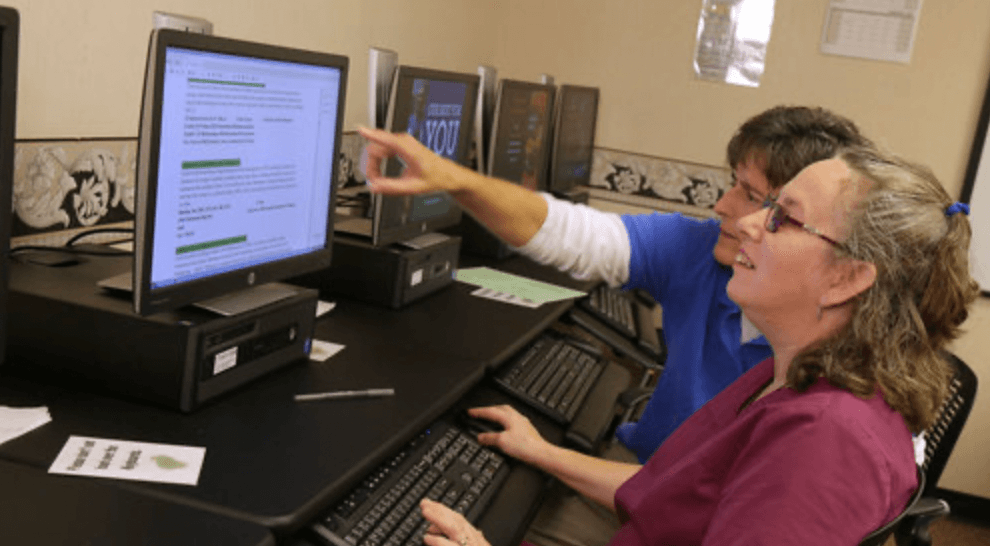 A man in a blue shirt and a woman in a dark pink shirt are looking at a computer screen. The man points to the screen as the woman sits in front of it.