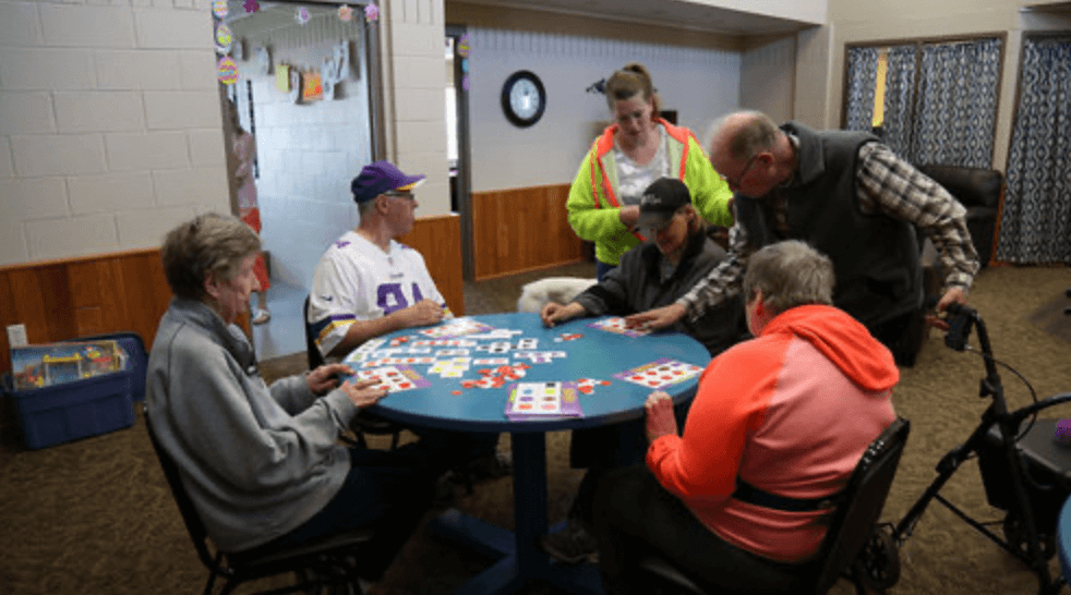 A group of five people are sitting around a round table playing cards. They are wearing grey, white, orange, and yellow shirts.
