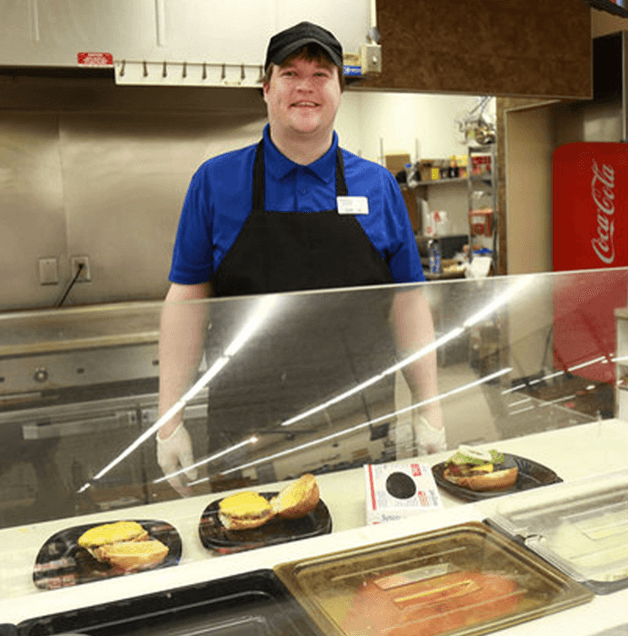 A man in a blue shirt and black apron stands behind a. Three differentfood lunch line. in front of him.