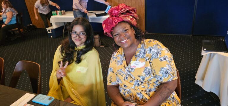Simi (right) and Reshim (left) smile are seated, leaning together and smiling. Reshim wheres a yellow sari and glasses. Simi wheres a red head wrap and floral top.
