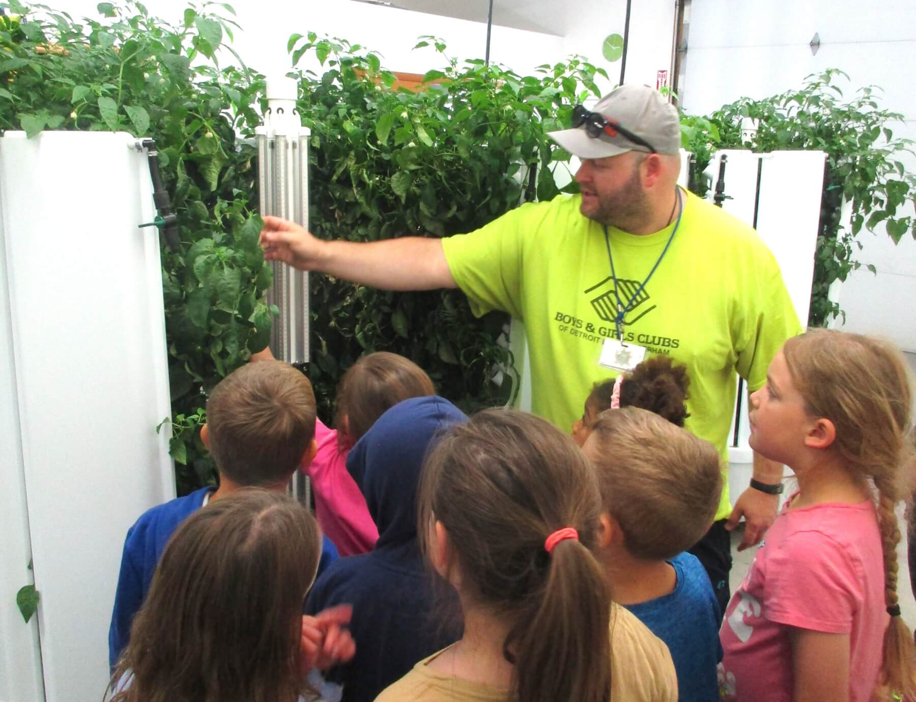 A man in a bright yellow Boys & Girls club shirt is showing a group of children plants in the hydroponic garden