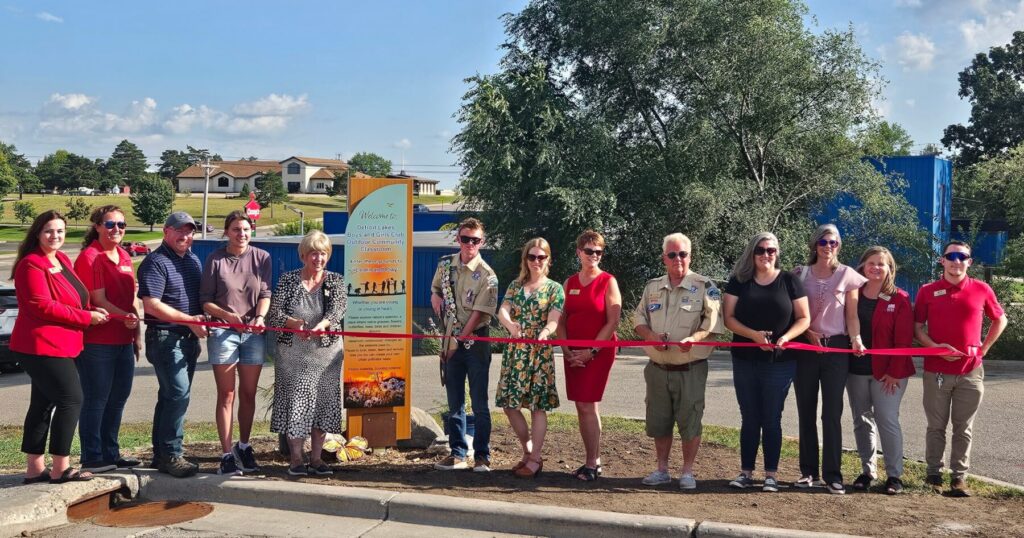 Pollinator Garden Ribbon cutting at Boys & Girls Club of Detroit Lakes. 13 people stand outside holding a long red ribbon and scissors.