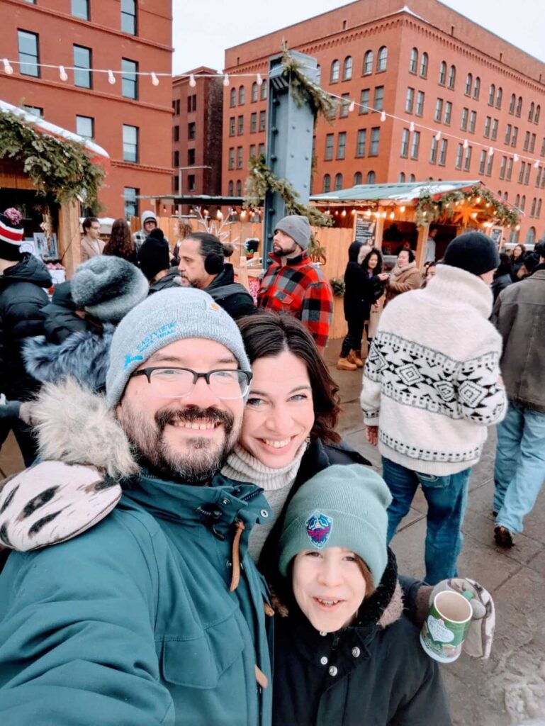 Jenna, Pete, and Max stand outside in a city in the winter. Pete and Max are wearing hats.