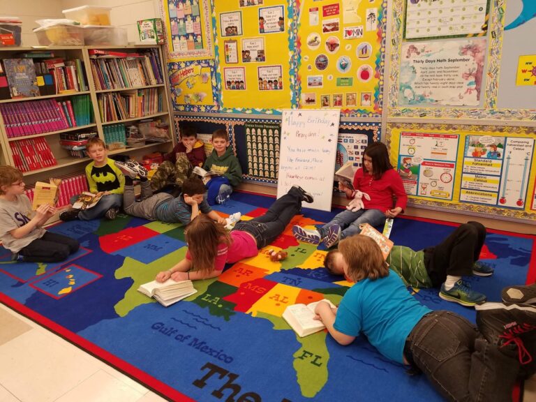 Elementary school students sitting and lying on a colorful U.S. map rug in a classroom, each reading a book. The walls are covered with bright educational posters and bulletin boards, and bookshelves filled with books line one side of the room.